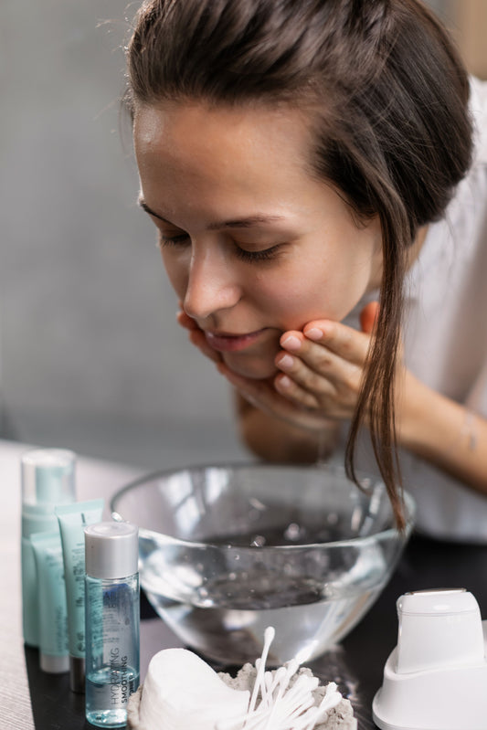 A young woman with dark hair rinses her face over a glass bowl of water, surrounded by aqua-toned skincare bottles, cotton pads, swabs and a cleansing device.