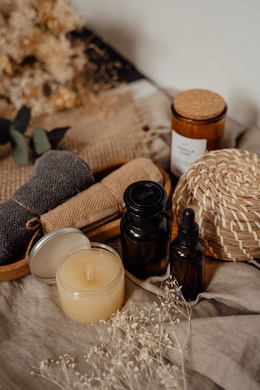 A cosy, natural skincare flat-lay: an open jar of golden scrub, amber apothecary bottles, a dropper serum, cork-lidded candle, woven basket, linen and dried flowers.