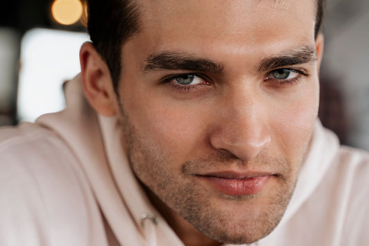 Close-up portrait of a young man with blue-grey eyes, thick dark brows, and light stubble, wearing a pale pink hoodie against a blurred, warm-lit background.