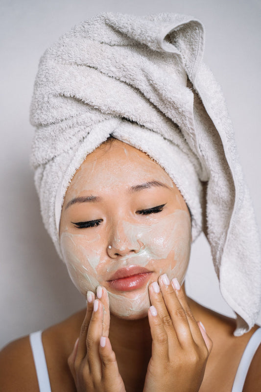 A young woman with a white towel wrapped around her hair applies a creamy, pale green face mask, eyes closed, against a soft grey background.