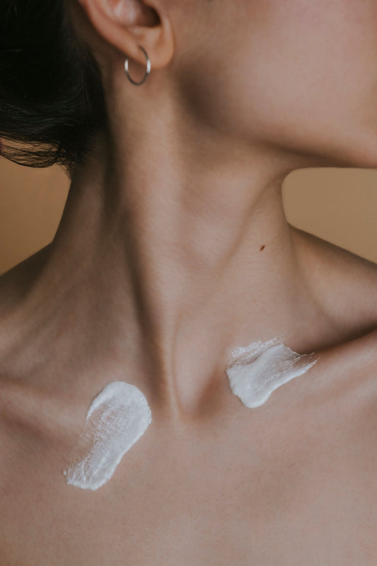 Close-up of a woman’s neck and collarbones, two smears of white cream on her skin, silver hoop earring, dark hair, soft warm beige background, minimalist.