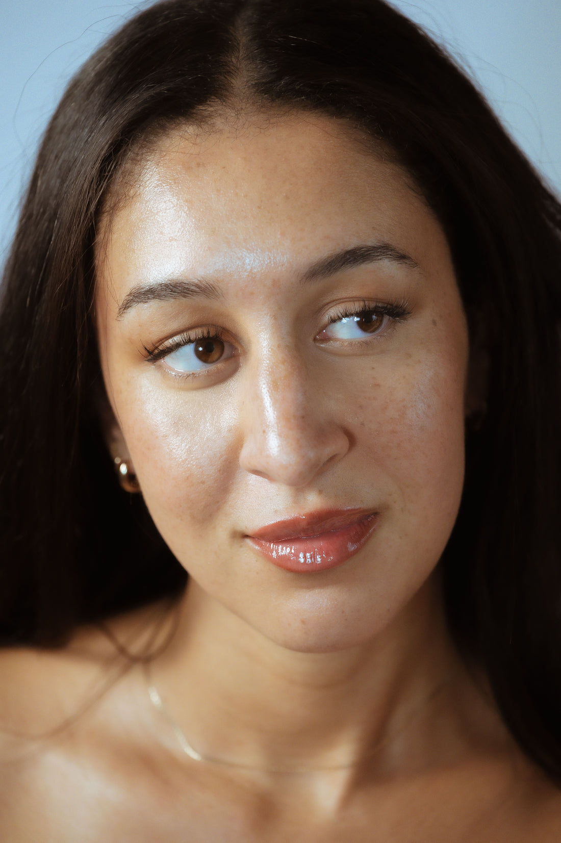 Close-up portrait of a woman with long dark hair, dewy freckled skin, brown eyes glancing sideways, glossy nude lips, gold earring, delicate necklace, soft-blue background.