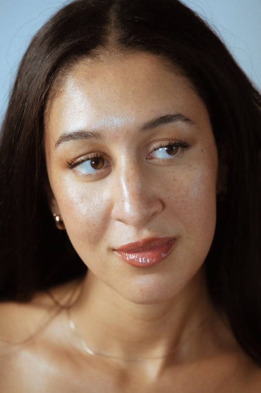 Close-up portrait of a woman with long dark hair, dewy freckled skin, brown eyes glancing sideways, glossy nude lips, gold earring, delicate necklace, soft-blue background.