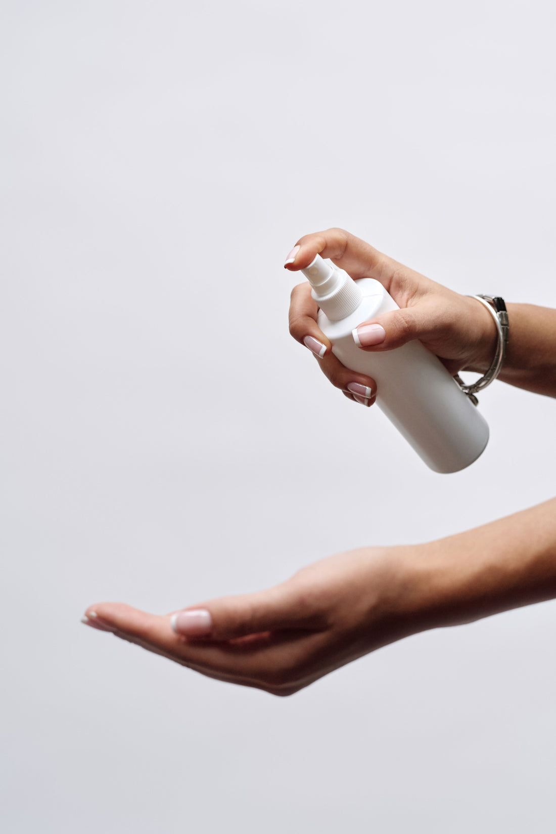 A manicured hand with a silver bracelet presses a white spray bottle, misting product onto an open, cupped palm below against a soft grey background.