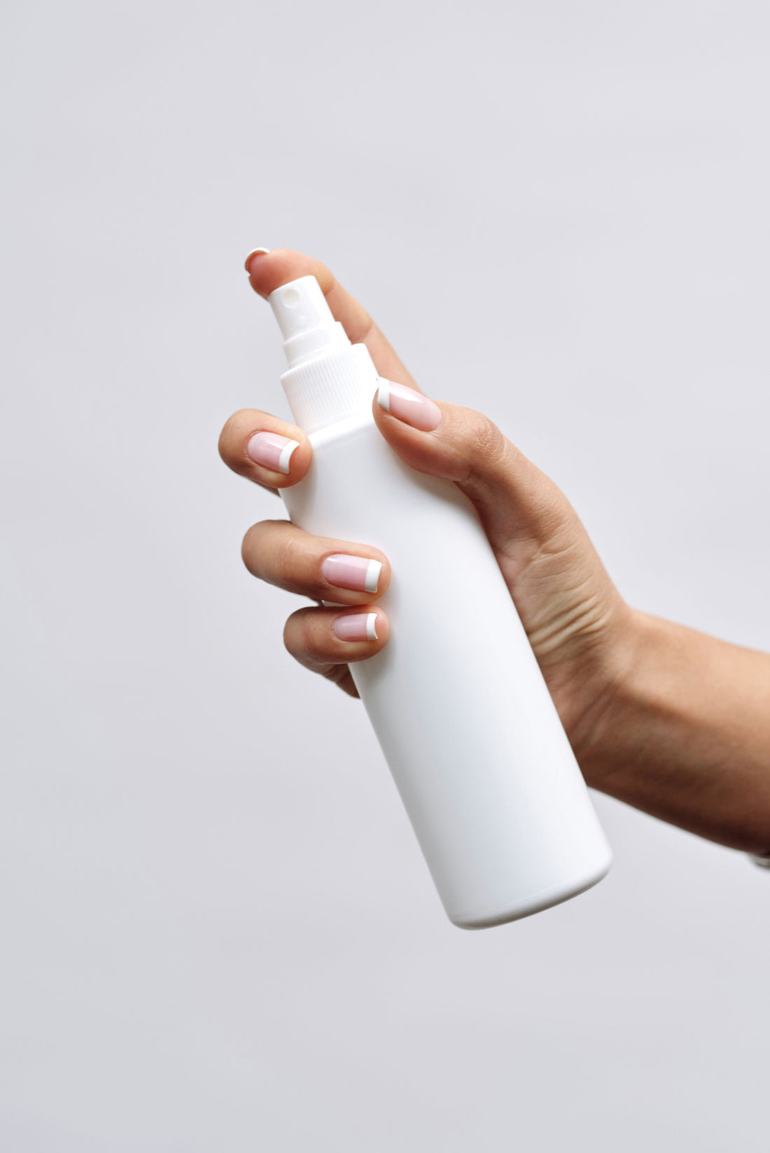 A hand with neat French-manicured nails holds a plain white spray bottle against a soft light-grey background, creating a clean, minimalist, unbranded skincare product aesthetic.