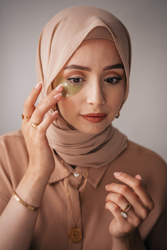 A young woman in a beige hijab and matching shirt applies a golden under-eye patch against a grey backdrop, wearing gold jewellery and terracotta matte lipstick.
