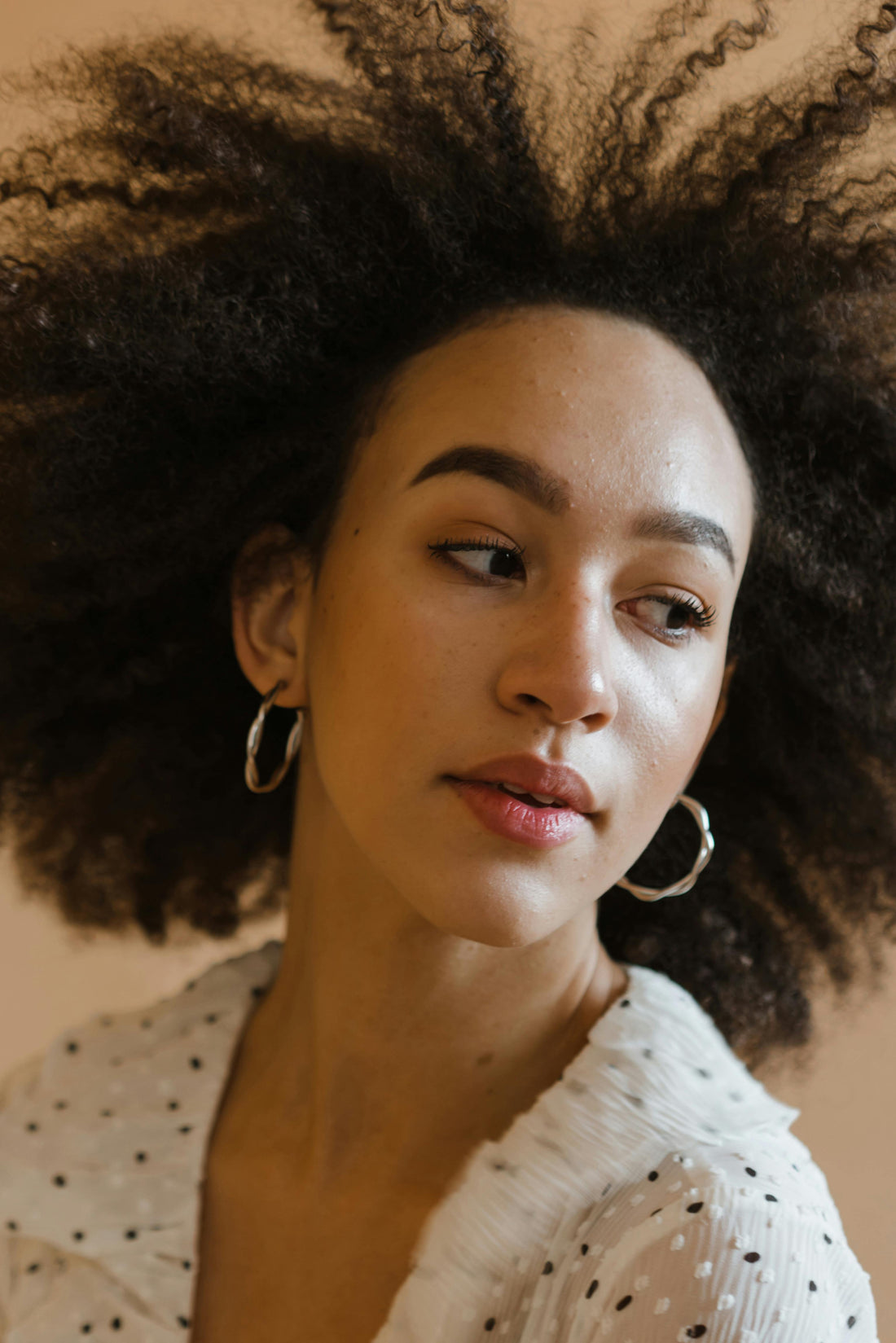 A close-up portrait of a woman with voluminous curly hair, warm skin, hoop earrings, and a sheer polka-dot blouse, gazing thoughtfully at the camera.
