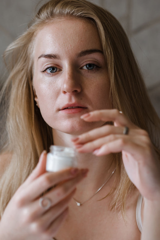 A close-up portrait shows a woman applying skincare cream, her natural face, blonde hair, delicate jewelry, and softly lit surroundings creating an intimate, candid moment.