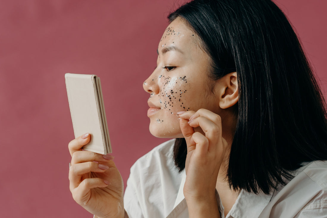 Left-sided portrait of a young Asian woman holding a mirror in her right hand, looking at a breakout on her face