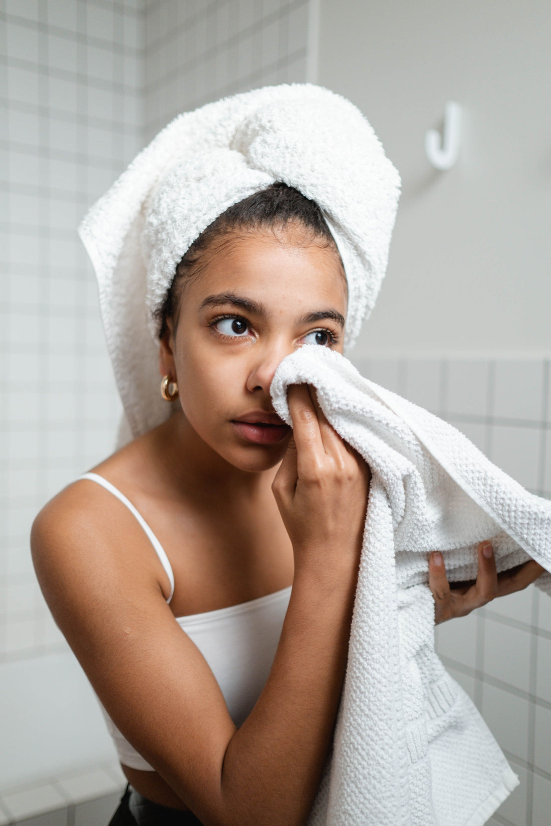 This is a head-and-shoulders photo of a young woman with her hair wrapped in a white towel, patting her face dry with another white towel in her right hand.
