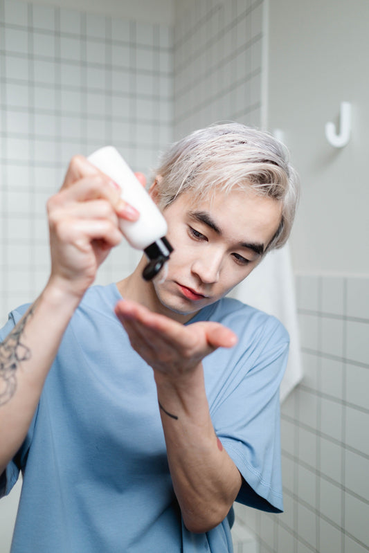A young man is squeezing the contents of a cylindrical bottle of a skincare product into his left hand, first placing it on the open palm of his right hand before applying it to his face.