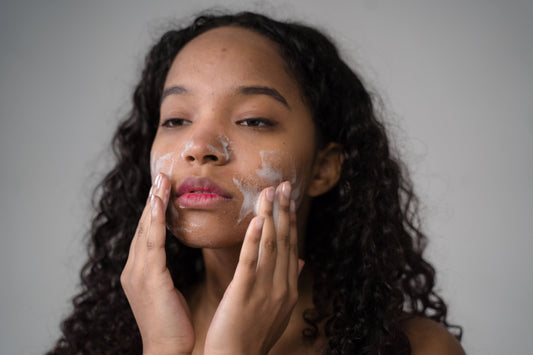 This is a picture of a dark-haired young woman of mixed-race heritage using both hands to lightly massage a cleansing cream into her face.