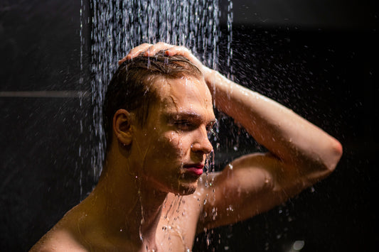 A young man with wet, slicked-back hair stands beneath a rainfall shower against a dark background, eyes half-closed, water streaming down his face and shoulders.