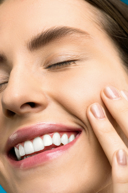 Close-up portrait of a smiling woman with closed eyes, clear skin, groomed eyebrows, glossy lips, white teeth, and fingers gently touching her cheek.