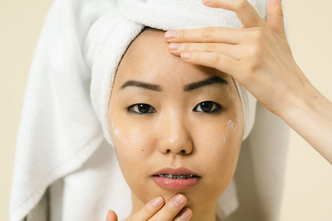 A young woman, hair wrapped in a white towel, massages cream into her face. She wears braces and gazes calmly against a soft beige background.
