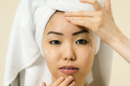 A young woman, hair wrapped in a white towel, massages cream into her face. She wears braces and gazes calmly against a soft beige background.