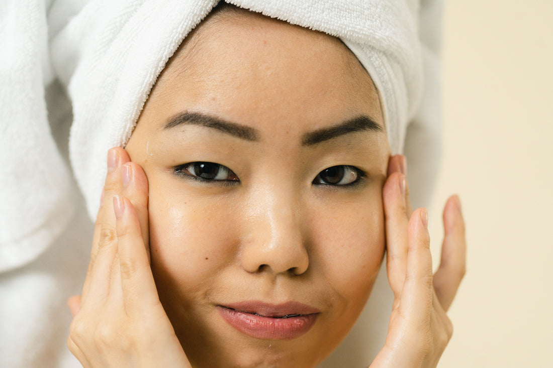 A young Asian woman with glowing skin is shown with her fingertips placed to the sides of her eyes
