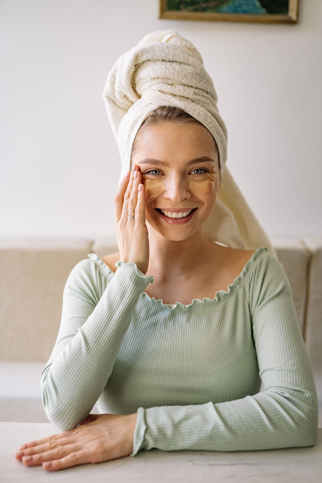A smiling woman with a cream towel wrapped around her hair wears golden under-eye patches and a sage ribbed top, seated at a minimalist table.