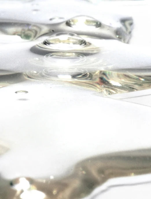 A close-up of clear, glossy liquid droplets and pools spreading across a white surface, catching soft light and reflections, evoking a fresh, hydrating serum texture.