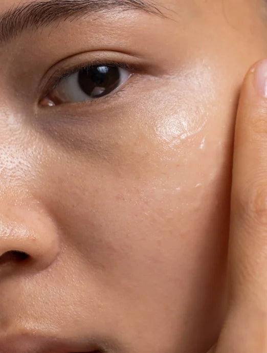 Extreme close-up of a woman’s face showing dewy, hydrated skin with visible pores, a glossy serum sheen on her cheek, and fingertips gently pressing nearby.