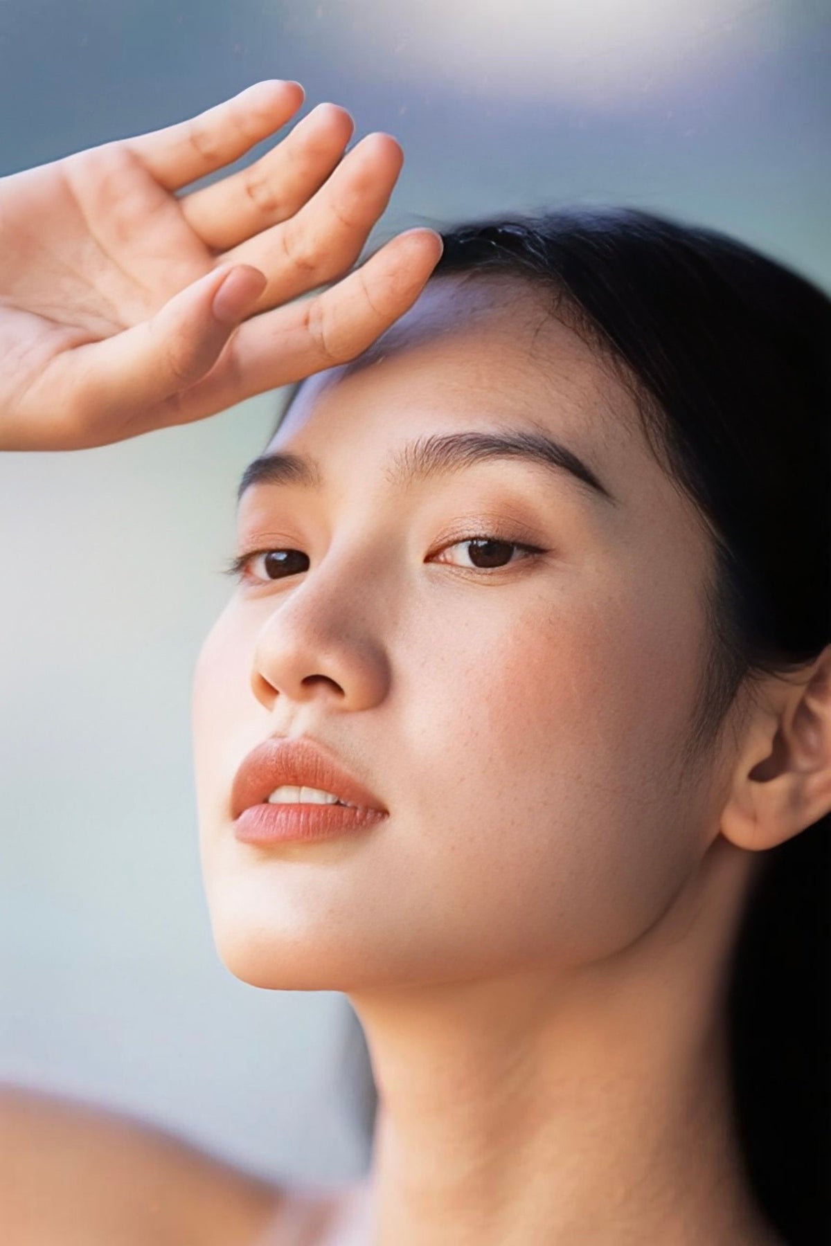 Close-up portrait of a woman with dark hair, looking upward, with her hand near her forehead against a soft light blue background.