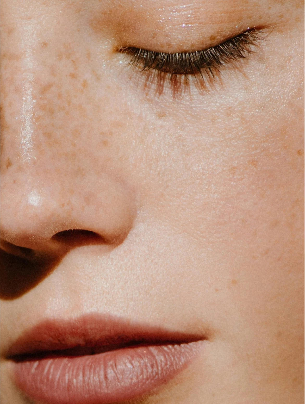 Intimate close-up of a freckled face with eyes closed, dewy glowing skin, natural pink lips, and warm light highlighting fresh, makeup-free texture and delicate lashes.